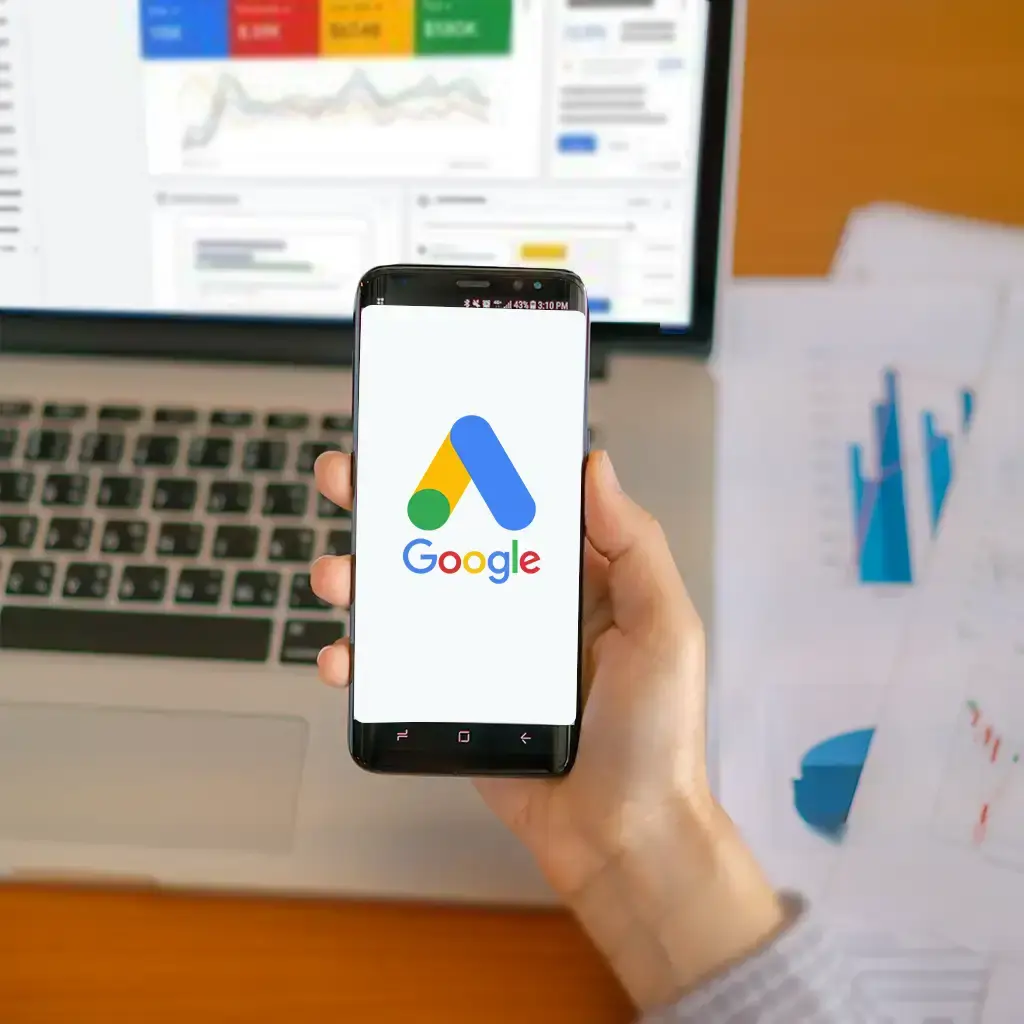 "Person holding a smartphone displaying the Google Ads logo in front of an open laptop showing ad performance dashboards and charts, with printed analytics reports on the desk.